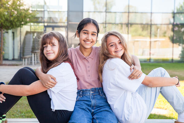 3 Mädchen sitzen draußen auf der Bank und umarmen sich.