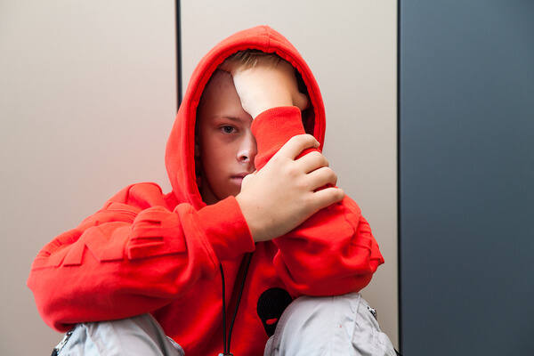 Ein  trauriger Junge sitz mit einem roten Kapuzenpullover am Gang und schaut in die Kamera.